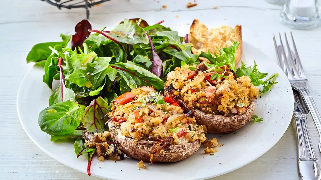 Gefüllte Champignons mit würzigem Couscous, Gemüse und Kräutern, serviert auf einem weißen Teller mit frischem Blattsalat und knusprigen Brotscheiben, angerichtet mit Besteck neben dem Teller. - Foto: LECKER @ Bauer Media Group Gefüllte Champignons mit würzigem Couscous, Gemüse und Kräutern, serviert auf einem weißen Teller mit frischem Blattsalat und knusprigen Brotscheiben, angerichtet mit Besteck neben dem Teller. - Foto: LECKER @ Bauer Media Group