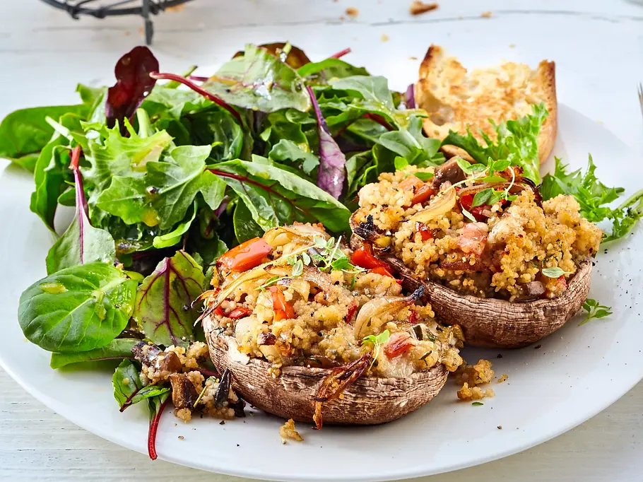Gefüllte Champignons mit würzigem Couscous, Gemüse und Kräutern, serviert auf einem weißen Teller mit frischem Blattsalat und knusprigen Brotscheiben, angerichtet mit Besteck neben dem Teller.