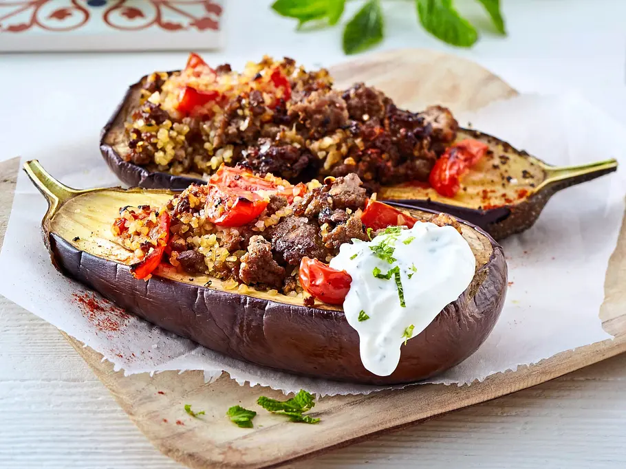 Gefüllte Auberginen mit Bulgur und Hackfleisch Zwei gefüllte Auberginenhälften mit einer Mischung aus Hackfleisch, Bulgur und Tomaten, serviert auf Backpapier auf einem Holzbrett, eine Hälfte mit einem Klecks Joghurt garniert.