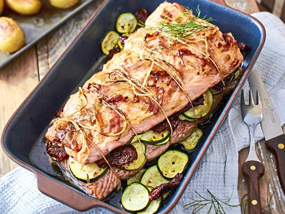 Ein mediterraner Lachsbraten, gebunden und goldbraun gegart, liegt auf einer Schicht aus Zucchini und getrockneten Tomaten in einer rechteckigen Auflaufform; daneben auf dem Holztisch Backblech mit kleinen gerösteten Kartoffeln.