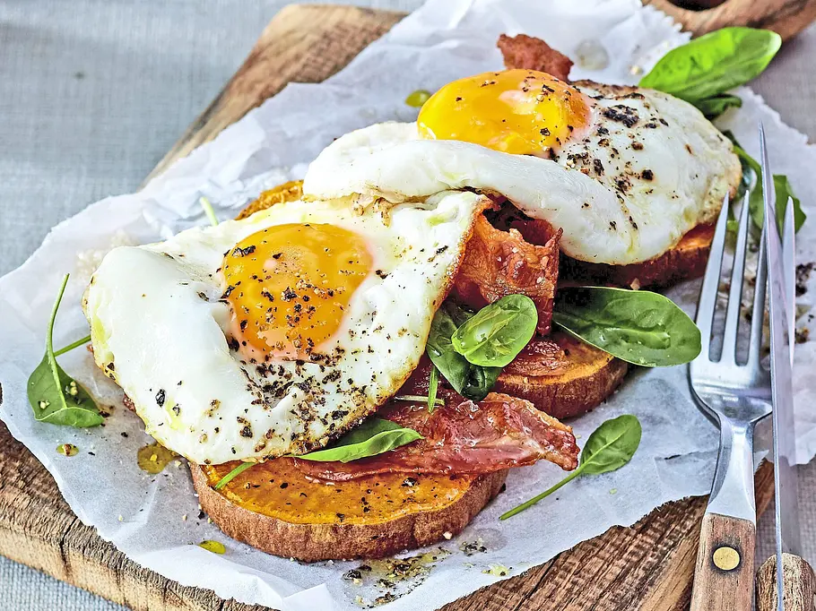 Scheiben von gebackener Süßkartoffel belegt mit kross gebratenem Bacon, Spinatblättern und zwei Spiegeleiern, angerichtet auf Backpapier und einem rustikalen Holzbrett.