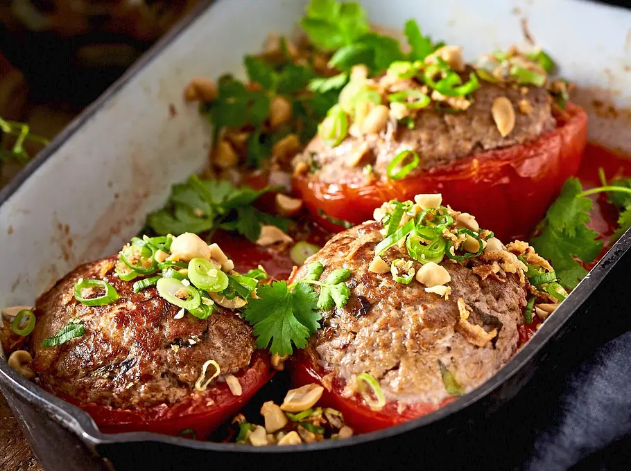 Drei große Tomaten, gefüllt mit würzigem Hackfleisch, in einer weißen Auflaufform, bestreut mit Frühlingszwiebeln, Erdnüssen und frischem Koriander.