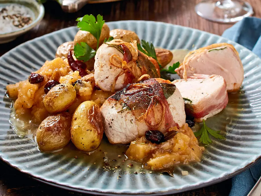 Zarte Puten-Medaillons, umwickelt mit Speck und Lorbeer, serviert mit kleinen Kartoffeln und fruchtigem Apfel-Kompott auf einem grünlich-blauen Keramikteller.