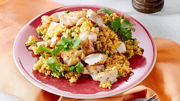 Zarte Fischfiletstücke auf lockerem Koriander-Couscous serviert, angerichtet auf einem rosafarbenen Teller mit passender Stoffserviette und Holzbesteck. - Foto: LECKER @ Bauer Media Group