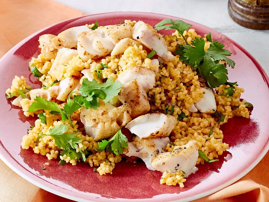 Zarte Fischfiletstücke auf lockerem Koriander-Couscous serviert, angerichtet auf einem rosafarbenen Teller mit passender Stoffserviette und Holzbesteck.