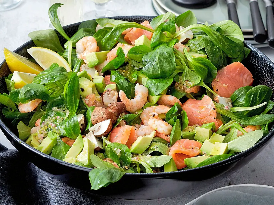 Frischer Salatmix mit Garnelen, Lachswürfeln, Avocado und Feldsalat in einer tiefen, schwarzen Schale, serviert mit Zitronenspalten.