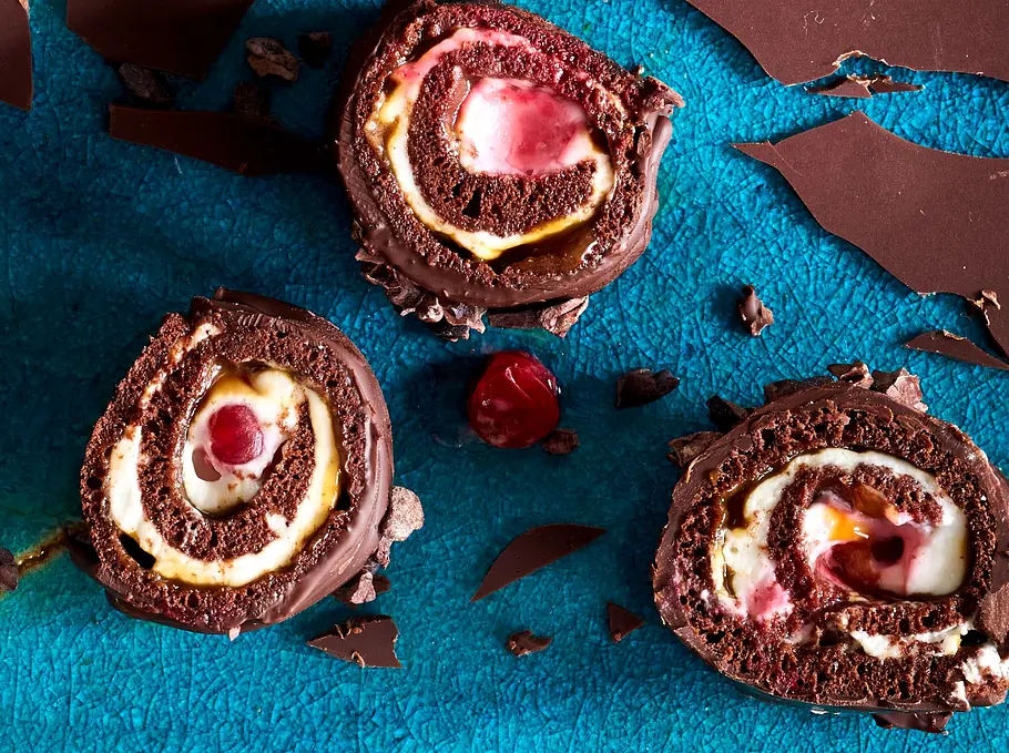 Schoko-Biskuitrollen mit heller Cheesecake-Füllung und roten Fruchtstücken, arrangiert auf einer türkisfarbenen Oberfläche mit zerbrochenen Schokoladenstücken.