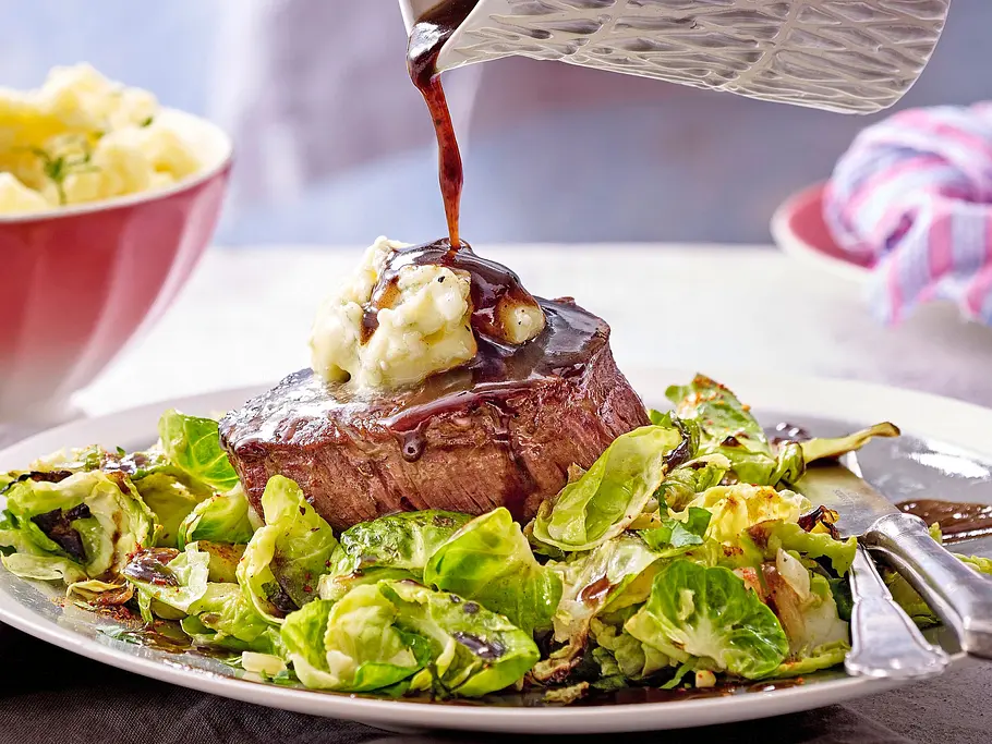 Zartes Rinderfilet mit einem Klecks würziger Butter, angerichtet auf gebratenen Rosenkohlblättern und mit dunkler Sauce beträufelt, serviert auf einem hellen Teller.