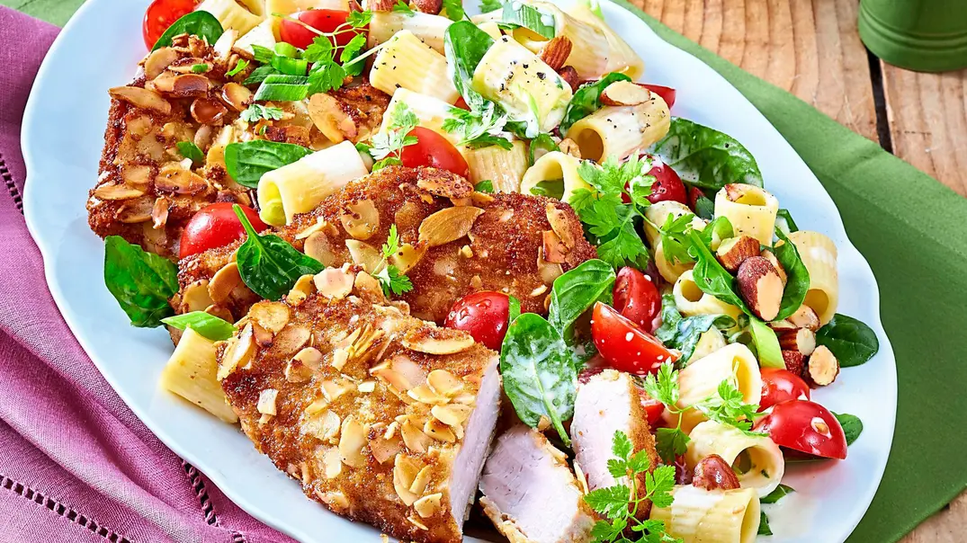 Knusprige Schnitzel mit Mandelkruste, angerichtet auf cremigem Nudelsalat mit Rigatoni, Cherrytomaten und frischem Blattspinat auf einer weißen Platte. - Foto: LECKER @ Bauer Media Group
