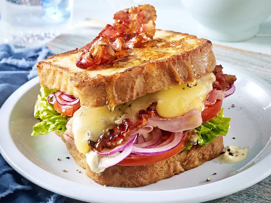 Herzhaftes Arme-Ritter-Sandwich aus goldbraun gebratenem Toast, gefüllt mit geschmolzenem Käse, knusprigem Bacon, Frischkäse, Salat, Tomaten und roten Zwiebeln, serviert auf einem weißen Teller.