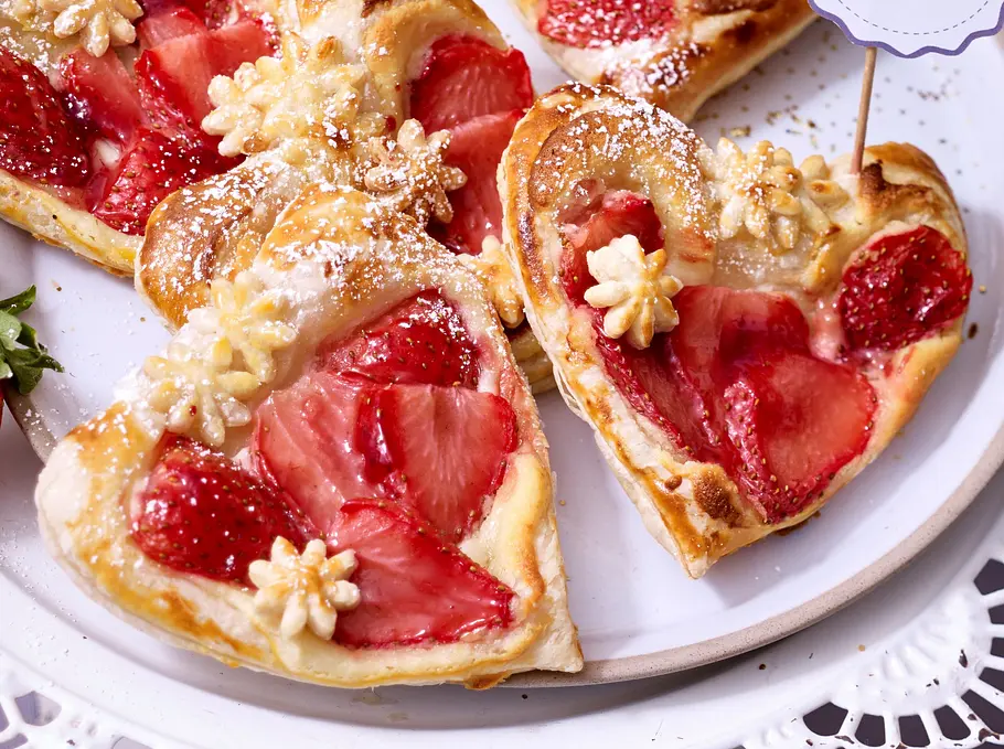 Blätterteig-Herzen mit Erdbeeren Rezept Blätterteig-Herzen mit frischen Erdbeerscheiben und Puderzucker liegen auf einer weißen Platte, daneben eine Erdbeere.