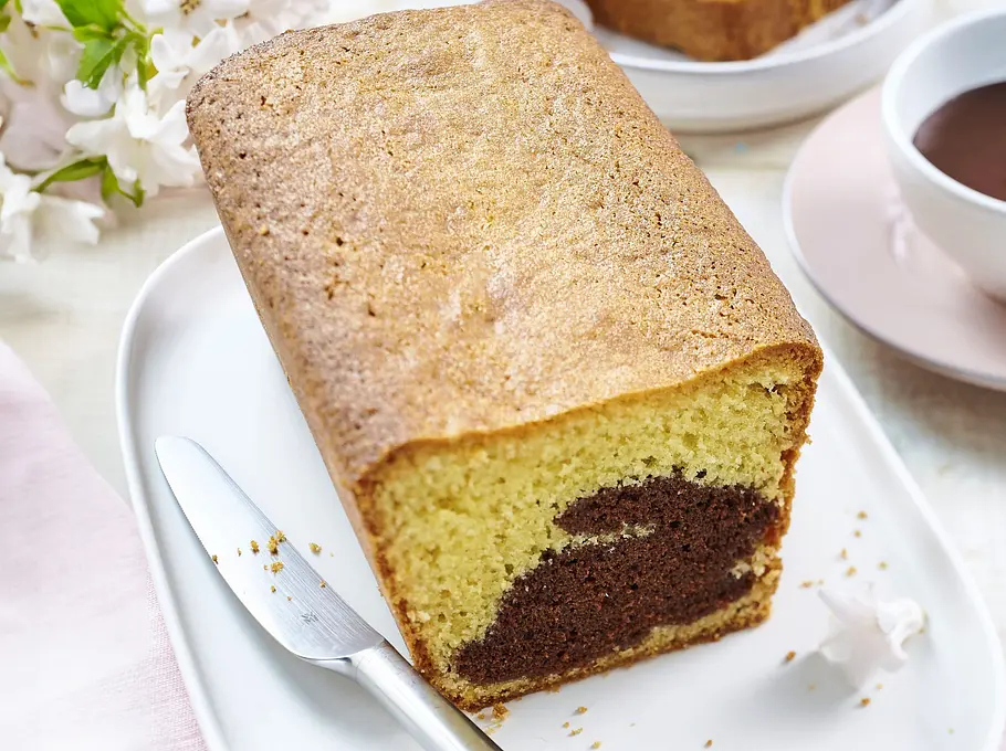 Brownie-Osterhase im Vanillekuchen-Versteck Rezept Saftiger Kastenkuchen aus Vanilleteig mit einem in der Mitte versteckten Brownie-Osterhasen, serviert auf einer weißen Platte mit einem Messer.