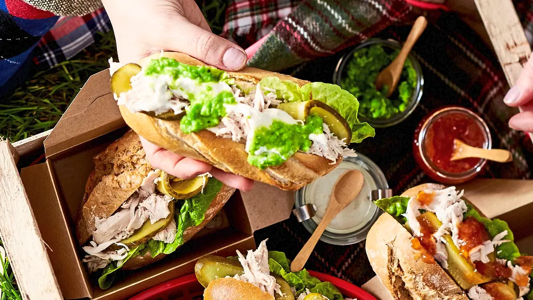 Saftige Pulled-Chicken-Sandwiches mit frischem Salat, eingelegten Gurken und cremigem Dressing in knusprigen Baguettebrötchen, serviert in kleinen Pappschalen auf einem Holztablett. - Foto: LECKER @ Bauer Media Group