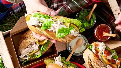 Saftige Pulled-Chicken-Sandwiches mit frischem Salat, eingelegten Gurken und cremigem Dressing in knusprigen Baguettebrötchen, serviert in kleinen Pappschalen auf einem Holztablett. - Foto: LECKER @ Bauer Media Group