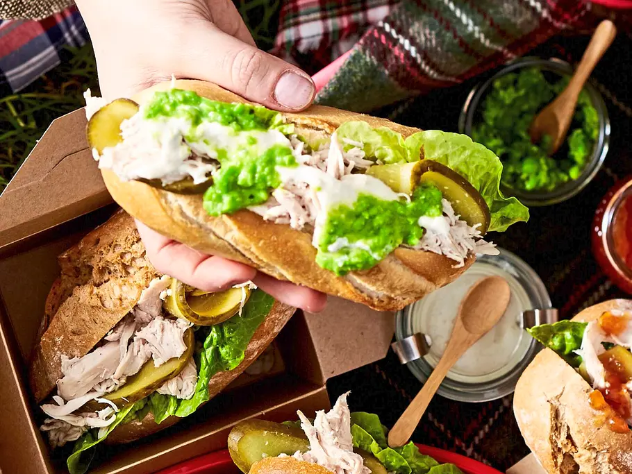 Pulled-Chicken-Sandwiches Saftige Pulled-Chicken-Sandwiches mit frischem Salat, eingelegten Gurken und cremigem Dressing in knusprigen Baguettebrötchen, serviert in kleinen Pappschalen auf einem Holztablett.