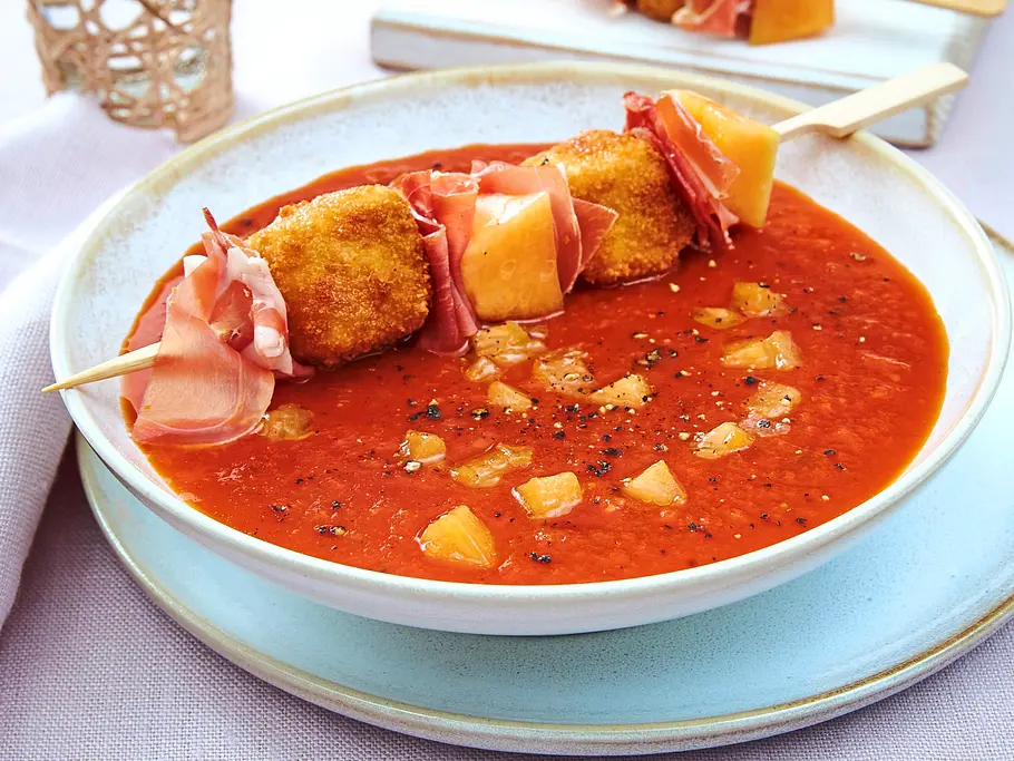 Tomaten-Melonen-Suppe in einer hellen Schale, serviert mit einem Holzspieß mit Melonenwürfeln, knusprig paniertem Käse und Serranoschinken, auf hellblauem Teller.