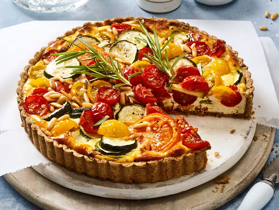 Tomaten-Ricotta-Tarte Herzhafte Tarte mit Ricotta, bunten Tomaten, Zucchinischeiben, Pinienkernen und frischem Rosmarin in einer runden Tarteform auf einem runden Holzbrett.