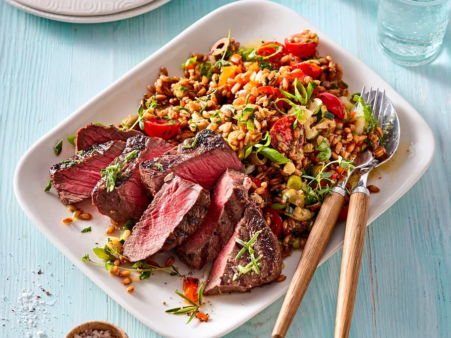 Dinkel-Gemüse-Salat mit Hüftsteak Saftige Hüftsteak-Scheiben auf einem Dinkel-Gemüse-Salat mit Tomaten und Kräutern, angerichtet auf einem weißen eckigen Teller auf hellblauer Holztischplatte.