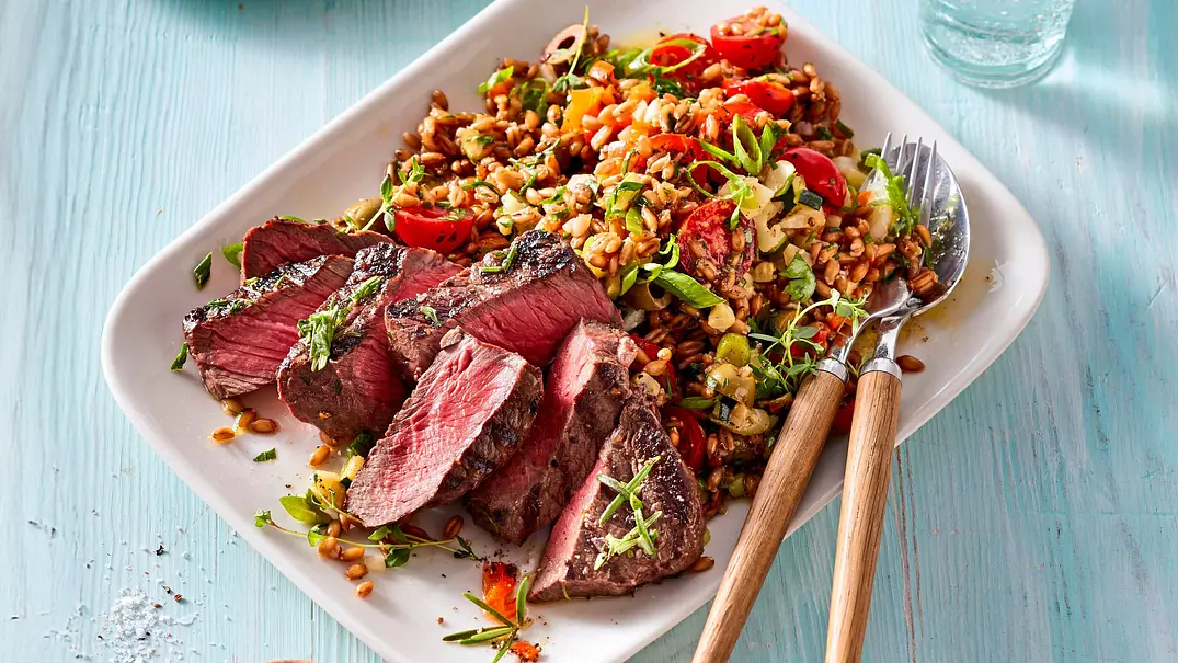 Saftige Hüftsteak-Scheiben auf einem Dinkel-Gemüse-Salat mit Tomaten und Kräutern, angerichtet auf einem weißen eckigen Teller auf hellblauer Holztischplatte. - Foto: LECKER @ Bauer Media Group