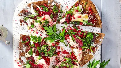 Falafel-Pizza in Stücke geschnitten, belegt mit frischem Gemüse, Roter Bete, Granatapfelkernen und Kräutern auf einem weißen, rechteckigen Teller. - Foto: LECKER @ Bauer Media Group