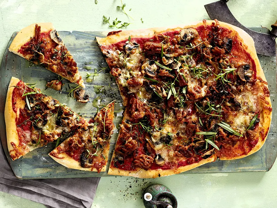 Bolognese-Pizza Rezept Herzhafte Bolognese-Pizza mit saftigem Hackfleisch, aromatischer Tomatensauce, geschmolzenem Käse und gebratenen Champignons, frisch gebacken auf knusprigem Pizzaboden, angerichtet auf einem rustikalen Holzbrett und mit grünen Kräutern bestreut.