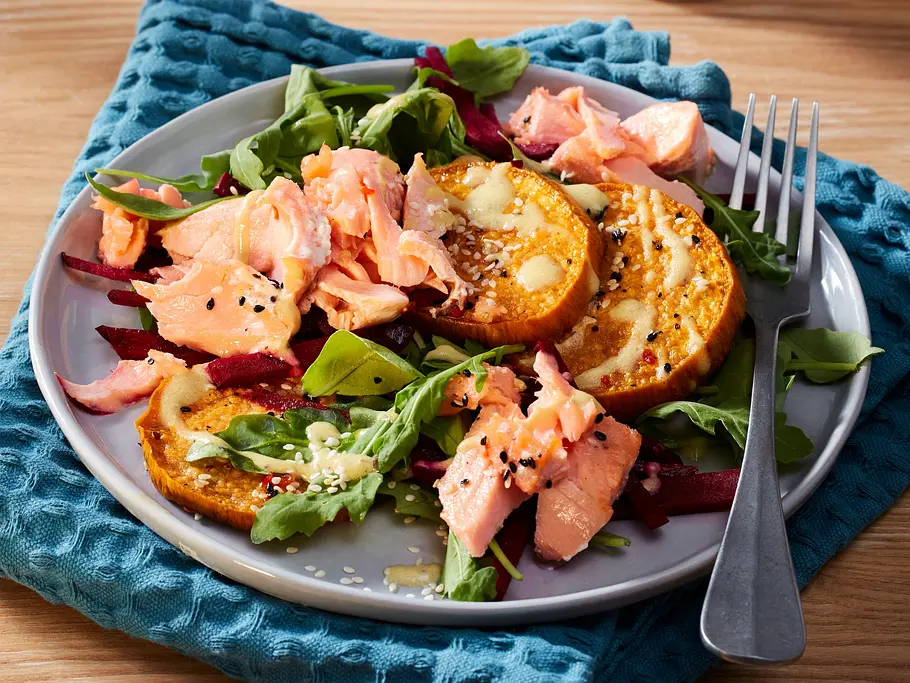Bunter Salat mit gerösteten Süßkartoffelscheiben, zarten Lachsstücken und frischem Blattgrün auf einem hellen Teller, der auf einer strukturierten, blauen Serviette liegt.