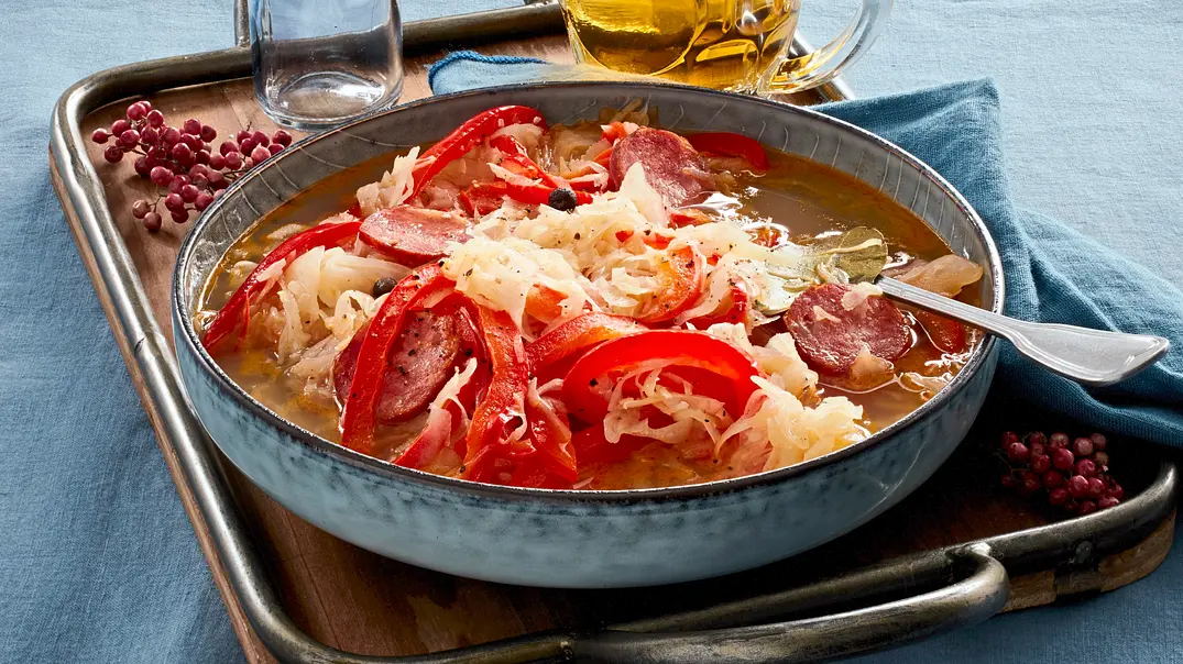 Herzhafter Eintopf mit Sauerkraut, Wurstscheiben und Paprikastreifen in einer rustikalen, blauen Schale auf einem Tablett, daneben ein Krug Bier und getrocknete Blüten als Dekoration auf einem blauen Tischtuch. - Foto: LECKER @ Bauer Media Group