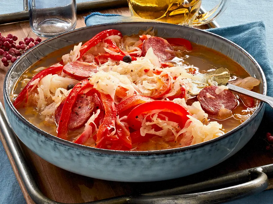 Herzhafter Eintopf mit Sauerkraut, Wurstscheiben und Paprikastreifen in einer rustikalen, blauen Schale auf einem Tablett, daneben ein Krug Bier und getrocknete Blüten als Dekoration auf einem blauen Tischtuch.