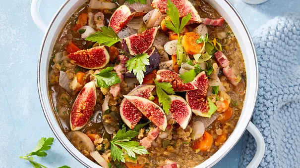Deftiger Linseneintopf im weißen Topf, garniert mit frischen Feigenstücken, Petersilie und Speckwürfeln, serviert auf hellblauer Unterlage mit gestricktem Tuch. - Foto: LECKER @ Bauer Media Group