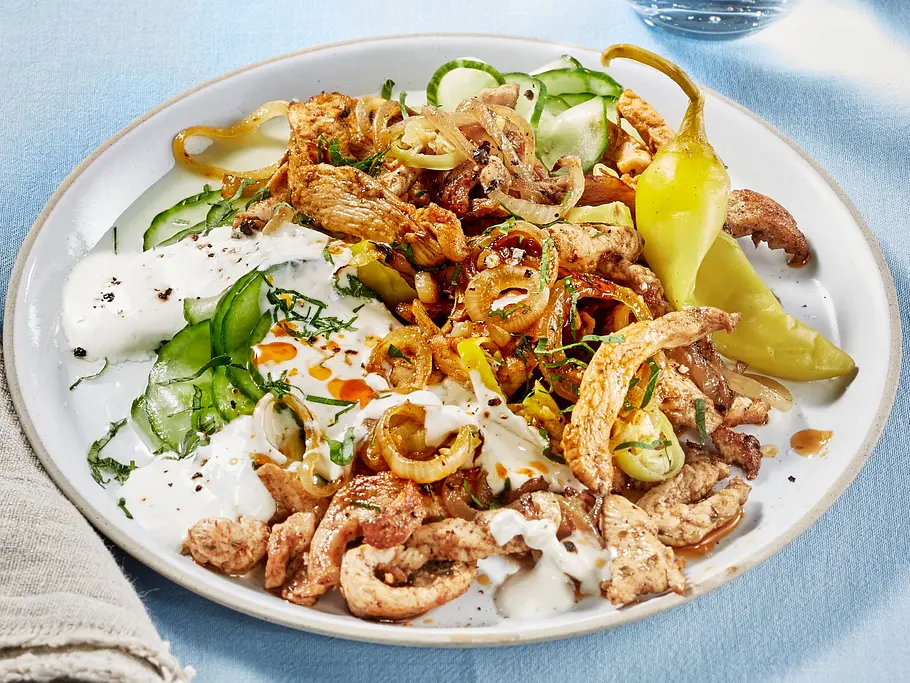 Zarte, gebratene Hähnchenstreifen mit Zwiebeln, serviert auf einem weißen, flachen Teller mit cremigem Joghurt-Dip, Gurkenscheiben und einer Peperoni, angerichtet auf einer hellblauen Stoffunterlage.