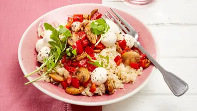 Blumenkohl-Risotto auf rosa Teller, belegt mit gebratenen Champignons, roter Paprika, Mini-Mozzarella und frischem Rucola, daneben Besteck auf rosafarbener Tischdecke. - Foto: LECKER @ Bauer Media Group