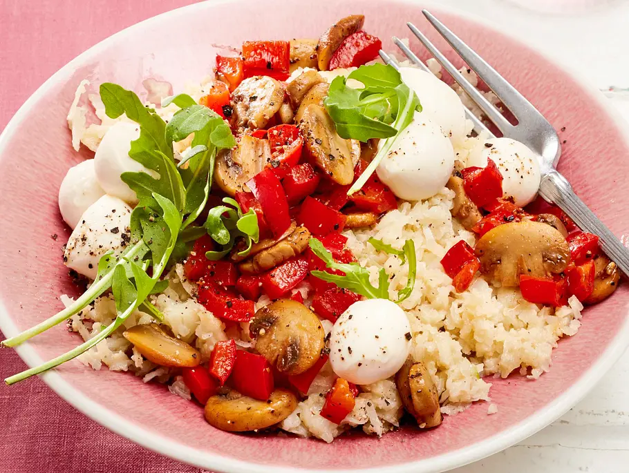 Blumenkohl-Risotto „Funghi e Mozzarella“ Rezept Blumenkohl-Risotto auf rosa Teller, belegt mit gebratenen Champignons, roter Paprika, Mini-Mozzarella und frischem Rucola, daneben Besteck auf rosafarbener Tischdecke.