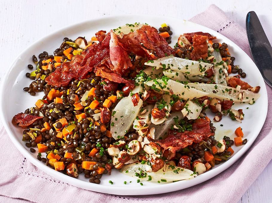 Salat aus schwarzen Linsen, Schwarzwurzeln und Karotten, bestreut mit gehackten Nüssen und Schnittlauch, serviert mit knusprigem Speck auf einem weißen Teller und rosafarbener Stoffserviette.