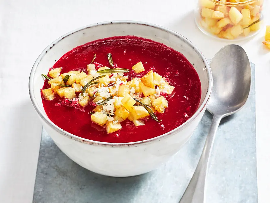 Rote-Bete-Suppe mit Kartoffel-Croûtons Eine Schale rote Suppe ist mit goldbraunen Würfeln und Kräutern garniert. Daneben liegen ein Löffel und ein Glas mit weiteren Würfeln.