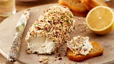 Rolle aus cremigem Ziegenkäse, mit gehackten Mandeln ummantelt, serviert auf einem runden Steinbrett mit knusprigen Brötchenchips. - Foto: LECKER @ Bauer Media Group