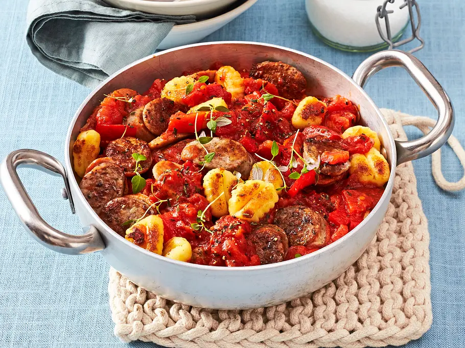 Herzhafter Eintopf aus goldgelb gebratenen Gnocchi, in Scheiben geschnittener Bratwurst und stückigen Tomaten, garniert mit frischen Kräutern in einer silbernen Kasserolle.