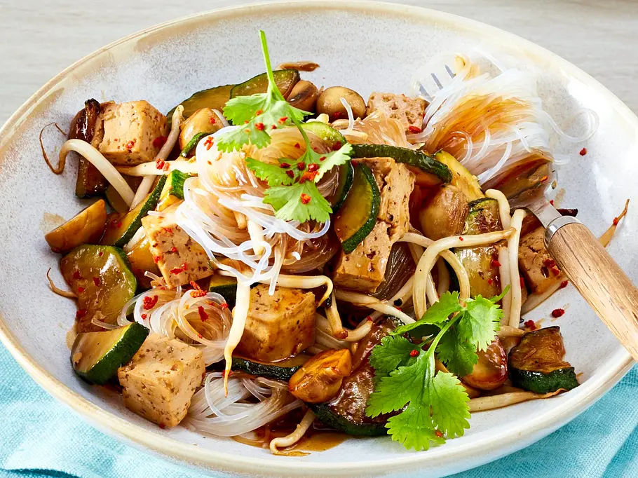 Ein Teller mit leichten Glasnudeln, gebratenem Tofu, Zucchini, Champignons und frischen Kräutern, alles in einer glänzenden Teriyaki-Sauce, angerichtet auf einer cremefarbenen Keramikschale mit hellem Holzbesteck und serviert auf einer hellblauen Stoffserviette.