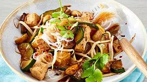 Ein Teller mit leichten Glasnudeln, gebratenem Tofu, Zucchini, Champignons und frischen Kräutern, alles in einer glänzenden Teriyaki-Sauce, angerichtet auf einer cremefarbenen Keramikschale mit hellem Holzbesteck und serviert auf einer hellblauen Stoffserviette. - Foto: LECKER @ Bauer Media Group Ein Teller mit leichten Glasnudeln, gebratenem Tofu, Zucchini, Champignons und frischen Kräutern, alles in einer glänzenden Teriyaki-Sauce, angerichtet auf einer cremefarbenen Keramikschale mit hellem Holzbesteck und serviert auf einer hellblauen Stoffserviette. - Foto: LECKER @ Bauer Media Group