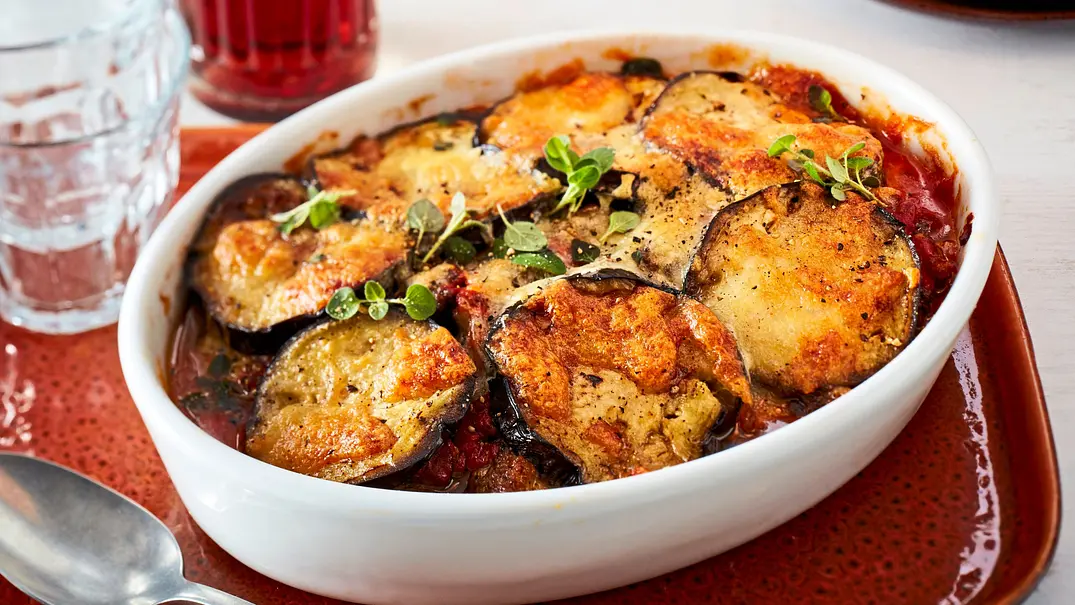 Überbackenes Auberginen-Linsen-Gratin in einer weißen Auflaufform auf einem roten Teller, mit goldbrauner Käseschicht und frischen Kräutern bestreut. Daneben ein silberner Löffel. - Foto: LECKER @ Bauer Media Group Überbackenes Auberginen-Linsen-Gratin in einer weißen Auflaufform auf einem roten Teller, mit goldbrauner Käseschicht und frischen Kräutern bestreut. Daneben ein silberner Löffel. - Foto: LECKER @ Bauer Media Group