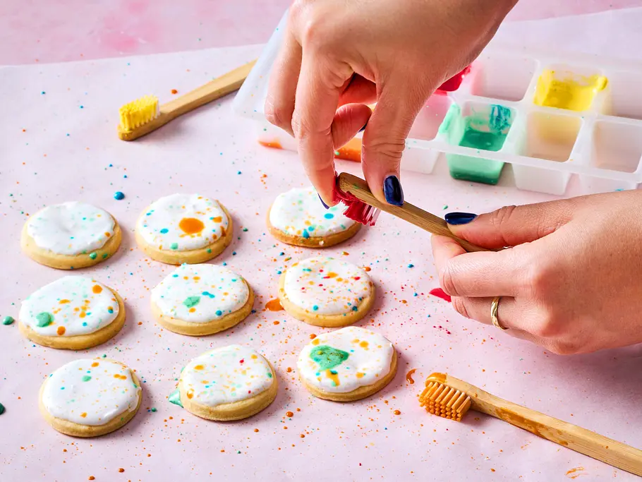 Farbkleckse mit Zahnbürsten auf Cookies verteilen Mit einer Zahnbürste wird bunter Zuckerguss auf die Plätzchen gespritzt.