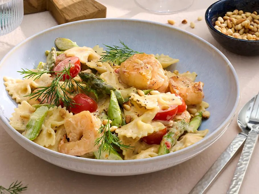 Farfalle-Pasta mit gebratenen Garnelen, grünem Spargel, Kirschtomaten und frischem Dill, angerichtet in einer hellen Schale. Daneben ein kleiner Teller mit gerösteten Pinienkernen.