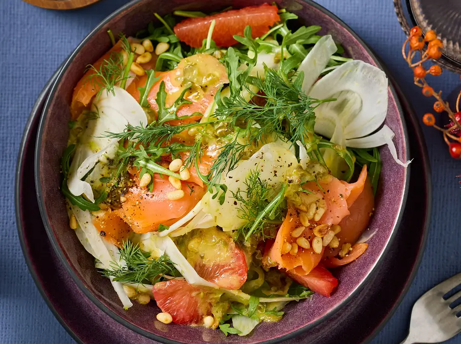Salat aus dünn geschnittenem Fenchel, Rucola, zarten Lachsscheiben und rosa Grapefruitfilets, bestreut mit Pinienkernen und frischem Dill, angerichtet in einer dunkelvioletten Schale.