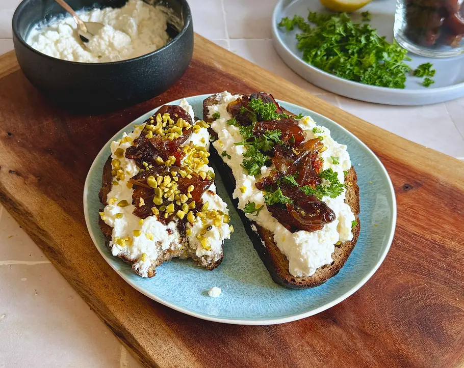 Feta-Stulle mit karamellisierten Datteln Rezept Zwei Scheiben Brot mit cremiger Fetacreme, karamellisierten Datteln, gehackten Pistazien und Petersilie auf einem Teller.