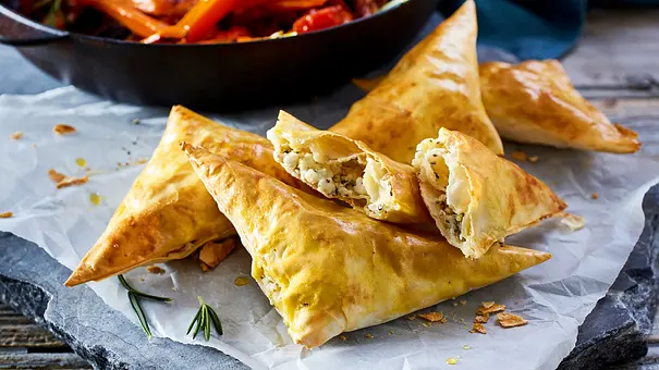Goldbraune dreieckige Teigtaschen mit heller Kräuterfüllung liegen auf Backpapier. Dahinter steht eine Pfanne mit Gemüse. - Foto: LECKER @ Bauer Media Group Goldbraune dreieckige Teigtaschen mit heller Kräuterfüllung liegen auf Backpapier. Dahinter steht eine Pfanne mit Gemüse. - Foto: LECKER @ Bauer Media Group