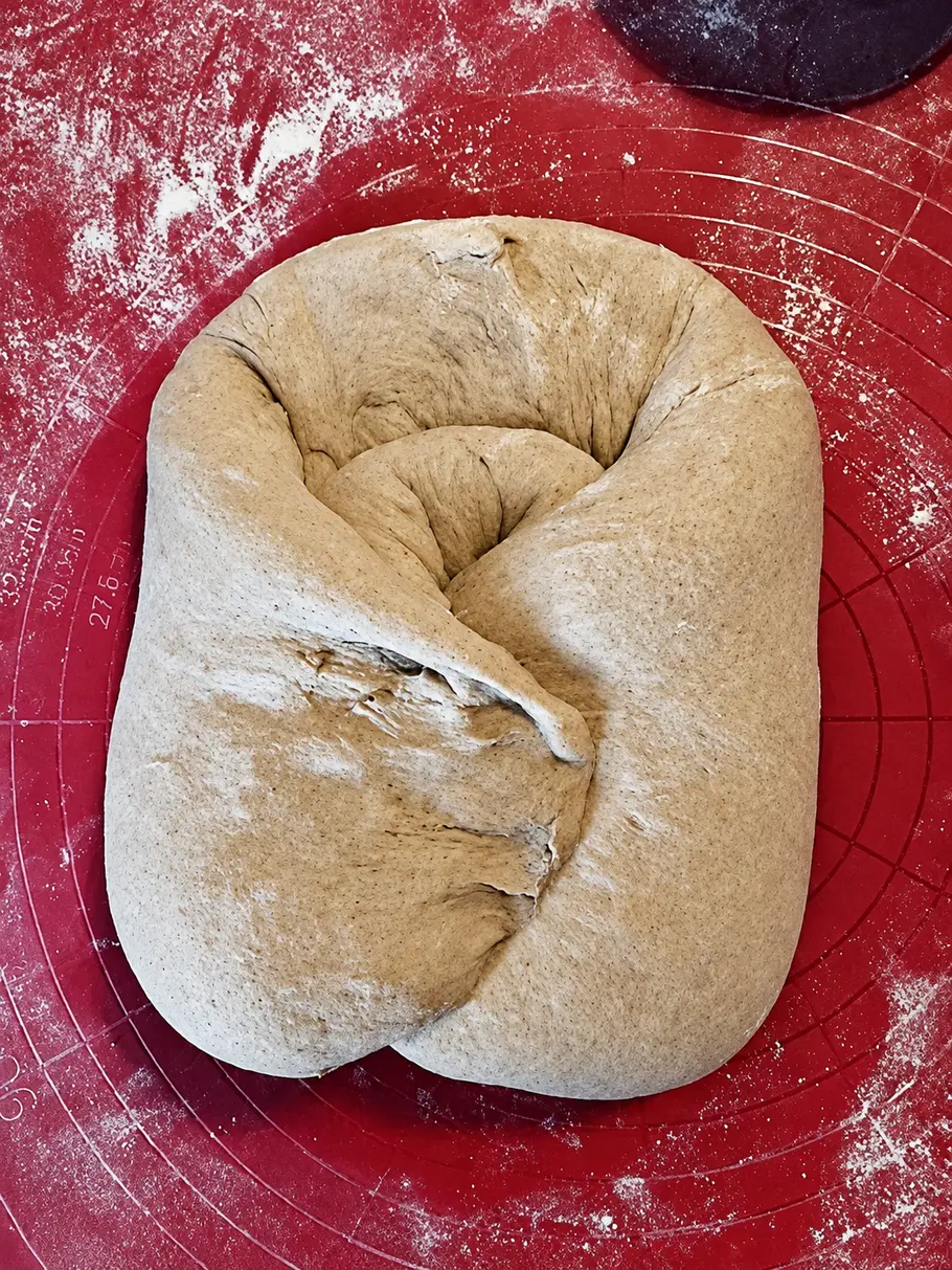 Final Shape Ein längliches Teigstück mit nach innen gefalteten Rändern liegt auf einer bemehlten roten Backmatte mit Kreismarkierungen.