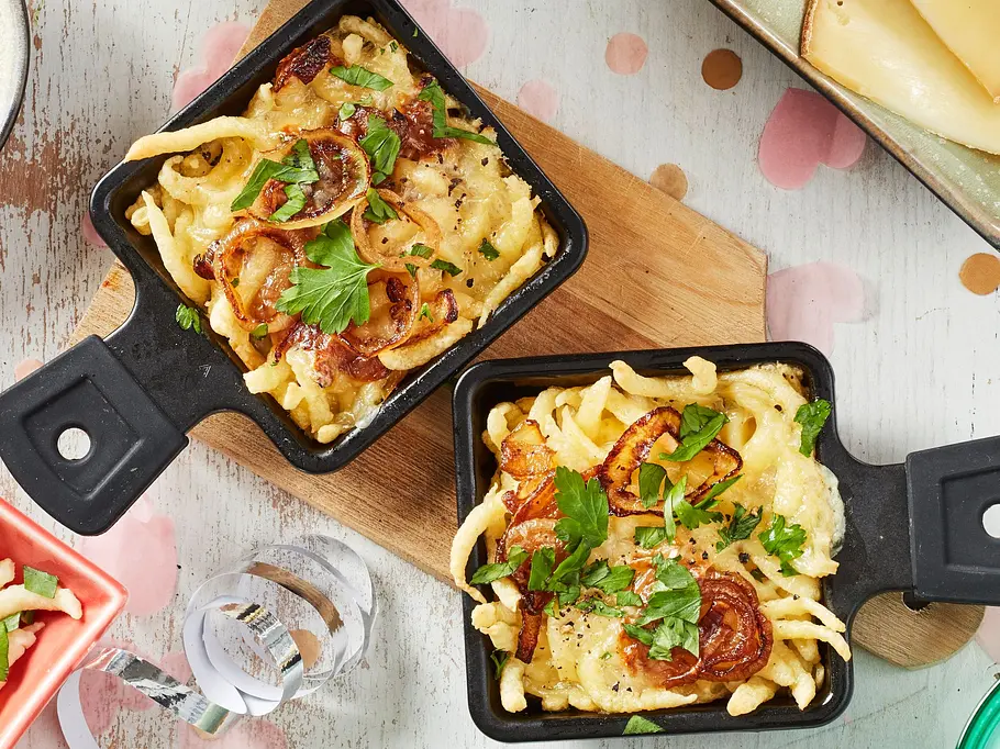 Frisch gebackene Käsespätzle in kleinen Raclettepfännchen, goldbraun überbacken mit geschmolzenem Käse und gebratenen Butter-Zwiebeln, serviert auf Keramiktellern und -schüsseln, garniert mit Petersilie.