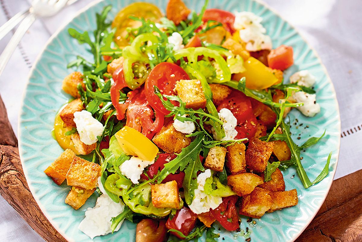 Ein bunter Salat mit Brotwürfeln, Rucola, Tomaten und Mozzarella auf einem blauen Teller.