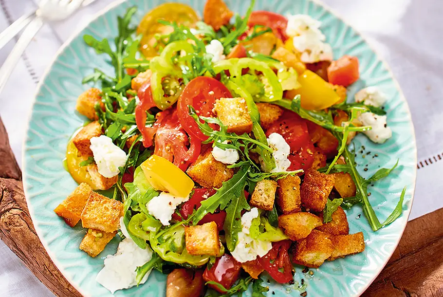 Brotsalat Ein bunter Salat mit Brotwürfeln, Rucola, Tomaten und Mozzarella auf einem blauen Teller.