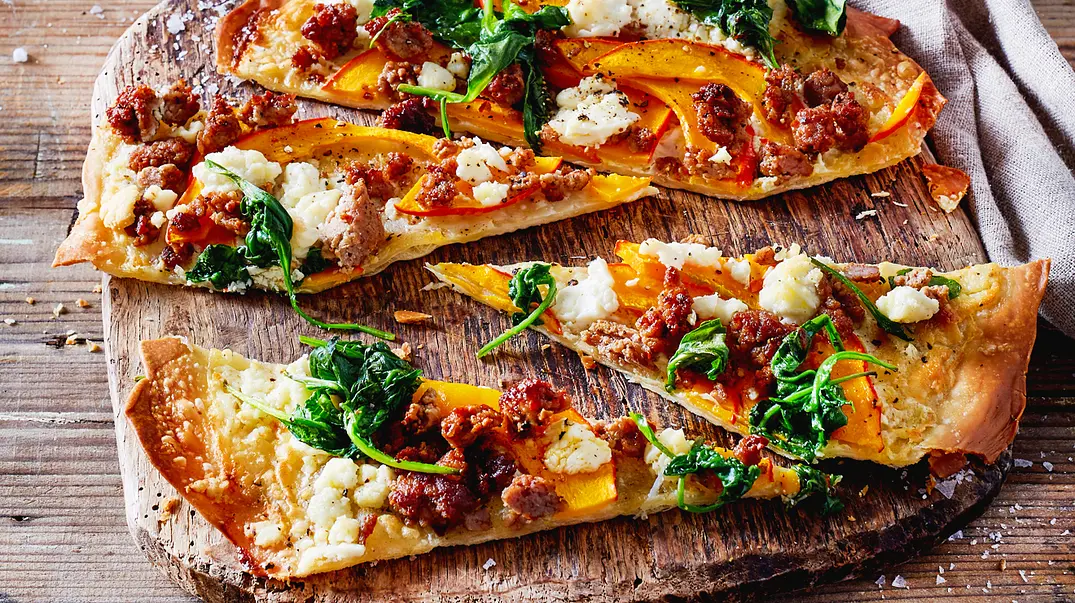 Auf einem Holzbrett liegen knusprige Flammkuchenstücke mit orangefarbenen Kürbisspalten, Blattspinat, Hackfleisch und weißem Käse. - Foto: ShowHeroes für LECKER @ Bauer Media Group Auf einem Holzbrett liegen knusprige Flammkuchenstücke mit orangefarbenen Kürbisspalten, Blattspinat, Hackfleisch und weißem Käse. - Foto: ShowHeroes für LECKER @ Bauer Media Group
