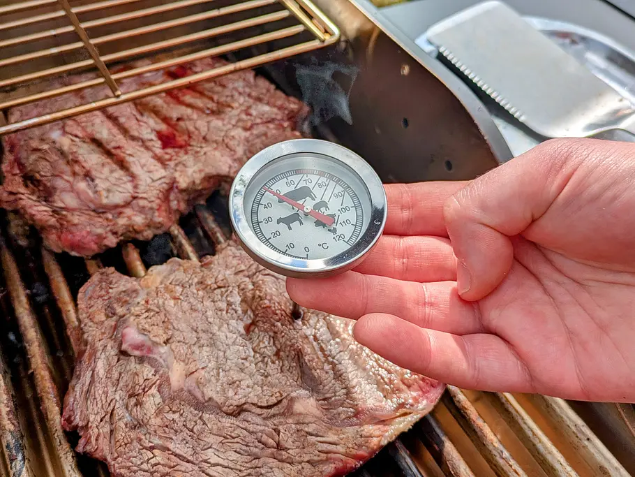Analoges Grillthermometer Zwei Steaks liegen auf einem Grillrost, eine Hand hält ein analoges Fleischtthermometer über dem Fleisch.